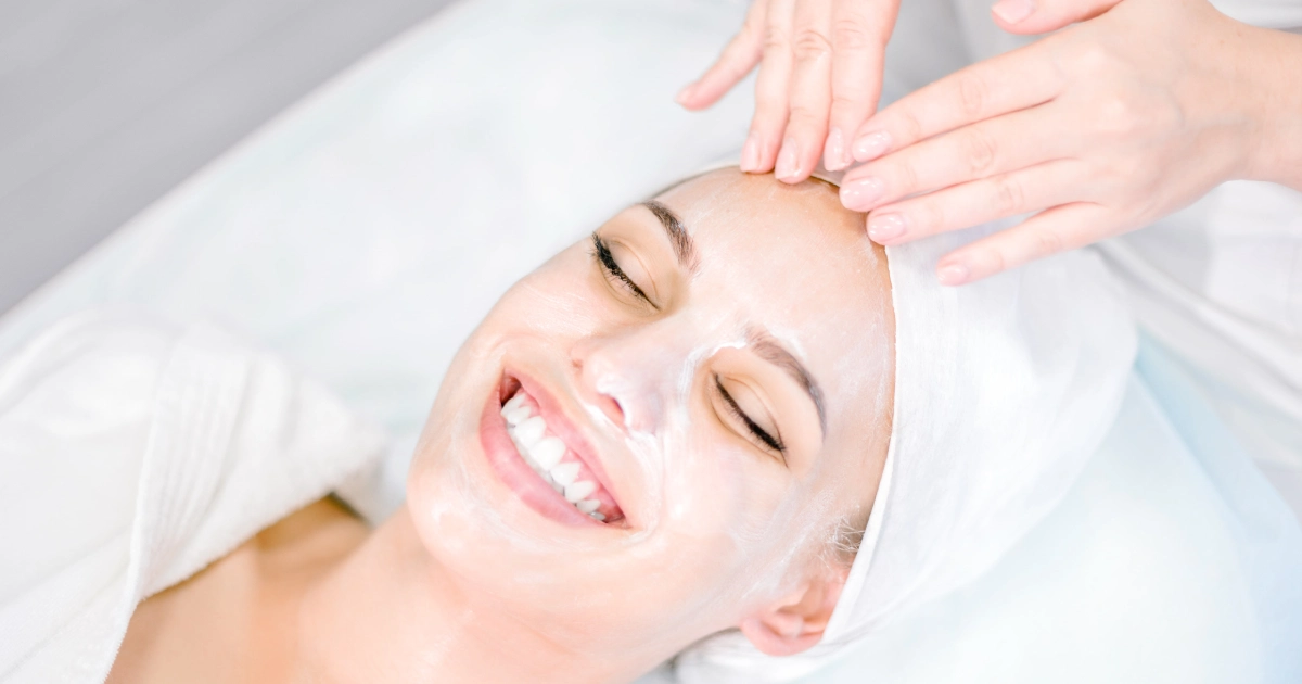 A smiling woman receives a Salmon Sperm Facial in Tampa, FL, with cream on her face and a towel around her head, while hands gently massage her forehead.